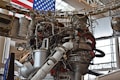 A complex assembly of machinery and pipes, possibly a rocket engine, is surrounded by various metal components and wiring. The background features part of an American flag, indicating a space or aerospace setting. Large, cylindrical objects and structural supports are visible alongside the intricate mechanical details.