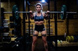 A woman is lifting a heavy barbell in a gym setting, showcasing tattoos on her legs and wearing athletic clothing. The gym environment is filled with weights and exercise equipment in the background, with dim lighting creating a focused, intense atmosphere.
