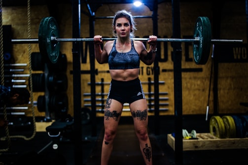 A woman is lifting a heavy barbell in a gym setting, showcasing tattoos on her legs and wearing athletic clothing. The gym environment is filled with weights and exercise equipment in the background, with dim lighting creating a focused, intense atmosphere.
