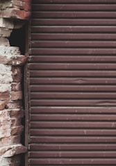 A close-up view of a partially exposed brick wall alongside a brown metal roll-up shutter. The bricks exhibit a mix of reddish and beige colors, and the surface appears weathered and aged. The metal shutter consists of horizontal slats with a slightly rough texture, indicating wear.