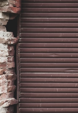 A close-up view of a partially exposed brick wall alongside a brown metal roll-up shutter. The bricks exhibit a mix of reddish and beige colors, and the surface appears weathered and aged. The metal shutter consists of horizontal slats with a slightly rough texture, indicating wear.