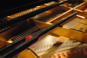A close-up of the interior of a grand piano, showing the strings, hammers, and dampers. The components are meticulously arranged, and the image accentuates the metallic strings and wooden hammers. The golden hue and warm lighting highlight the intricate details and craftsmanship of the piano.