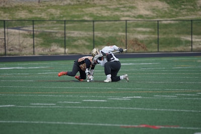 Two athletes wearing protective gear and holding lacrosse sticks face off on a sports field. The grass is marked with white lines, and the background features a fence and a grassy area.