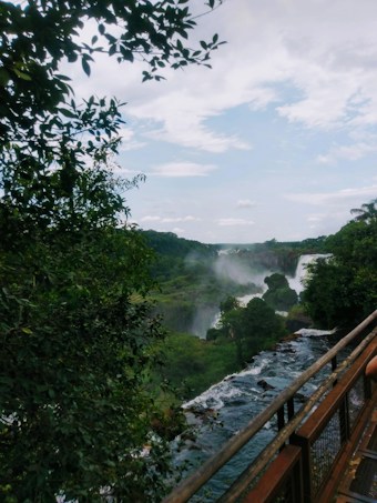 A lush green landscape with dense foliage surrounding a cascading waterfall. The waterfall flows alongside a wooden walkway, bordered by metal railings, suggestive of a nature trail for visitors. Clouds partly cover the sky, adding to the scenic beauty.