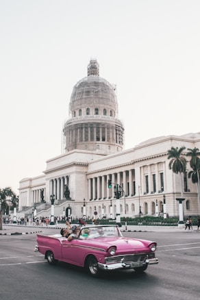A classic pink convertible car is driving past a large, neoclassical building with a dome under renovation. The street is busy with pedestrians, and palm trees are visible, suggesting a warm location. The building features grand columns and detailed architectural elements.