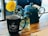 A dark coffee mug with 'SIP JAVA CO.' printed on it sits on a wooden table beside a small vase with a bright yellow flower. Next to them is a blue teapot, accompanied by a piece of paper with a spiral edge.