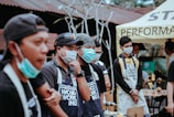 Several people wearing masks and aprons stand in a group at an outdoor event, likely related to woodworking. They appear to be focused and attentive, possibly listening to a speaker or demonstration. The setting includes a tent and various tools and equipment in the background.