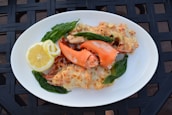 A white plate holds a serving of baked lobster with melted cheese, garnished with fresh basil leaves. A slice of lemon is placed on the side for added zest. The dish is presented on a textured black surface.