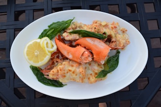 A white plate holds a serving of baked lobster with melted cheese, garnished with fresh basil leaves. A slice of lemon is placed on the side for added zest. The dish is presented on a textured black surface.