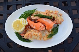 A white plate holds a serving of baked lobster with melted cheese, garnished with fresh basil leaves. A slice of lemon is placed on the side for added zest. The dish is presented on a textured black surface.