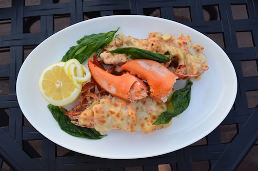 A white plate holds a serving of baked lobster with melted cheese, garnished with fresh basil leaves. A slice of lemon is placed on the side for added zest. The dish is presented on a textured black surface.