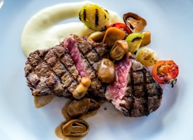 A well-cooked steak is garnished with mushrooms, grilled cherry tomatoes, and charred onions, surrounded by a creamy, white sauce. The steak shows clear grill marks, indicating it was cooked on a grill.