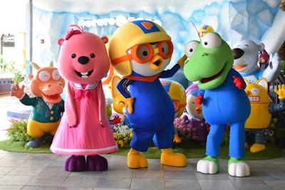 Several colorful cartoon character statues are grouped together. There is a pink bear wearing a dress, a yellow bird with orange glasses and a helmet, and a green amphibian-like creature. They are surrounded by additional characters including a smiling bear and a robot in colorful environments with a backdrop of ice or snow.