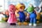 Several colorful cartoon character statues are grouped together. There is a pink bear wearing a dress, a yellow bird with orange glasses and a helmet, and a green amphibian-like creature. They are surrounded by additional characters including a smiling bear and a robot in colorful environments with a backdrop of ice or snow.