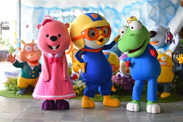 Several colorful cartoon character statues are grouped together. There is a pink bear wearing a dress, a yellow bird with orange glasses and a helmet, and a green amphibian-like creature. They are surrounded by additional characters including a smiling bear and a robot in colorful environments with a backdrop of ice or snow.