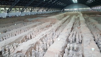 A vast archaeological site houses numerous life-sized statues of soldiers and horses, organized in rows within a large covered excavation pit. The figures are intricately detailed and arranged as if in a military formation. There is a viewing area along the perimeter where visitors are observing the display.