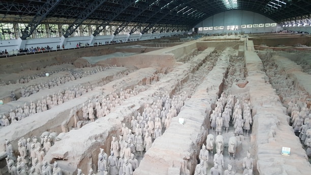 A vast archaeological site houses numerous life-sized statues of soldiers and horses, organized in rows within a large covered excavation pit. The figures are intricately detailed and arranged as if in a military formation. There is a viewing area along the perimeter where visitors are observing the display.