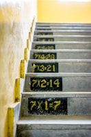 A staircase with multiplication tables painted on each step in bold yellow numbers against a black background. The walls are a light beige, and the steps are concrete.