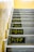 A staircase with multiplication tables painted on each step in bold yellow numbers against a black background. The walls are a light beige, and the steps are concrete.