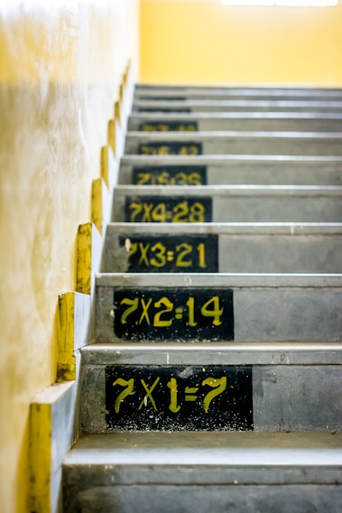A staircase with multiplication tables painted on each step in bold yellow numbers against a black background. The walls are a light beige, and the steps are concrete.