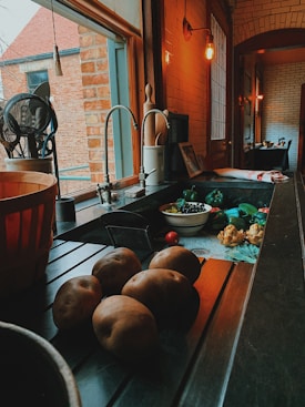 A cozy kitchen counter with various vegetables including potatoes, artichokes, and a bowl of blueberries. The scene is warmly lit with an overhead bulb, casting soft shadows. A large window provides a view to an adjacent brick building, and kitchen utensils are neatly arranged by the sink.
