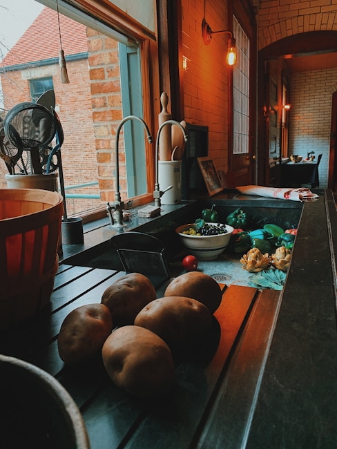 A cozy kitchen counter with various vegetables including potatoes, artichokes, and a bowl of blueberries. The scene is warmly lit with an overhead bulb, casting soft shadows. A large window provides a view to an adjacent brick building, and kitchen utensils are neatly arranged by the sink.