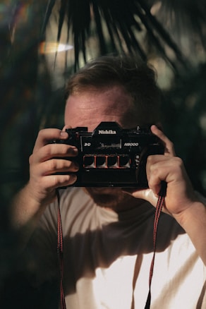 A person holds up a Nishika N8000 3D camera, partially obscuring their face. The setting is outdoors with dappled sunlight filtering through leaves, creating a play of light and shadow.