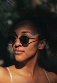 A person with curly hair is wearing dark sunglasses and has a soft smile. A rainbow light effect is seen across the person's forehead. The background appears to be outdoors with a hint of foliage and dappled sunlight.