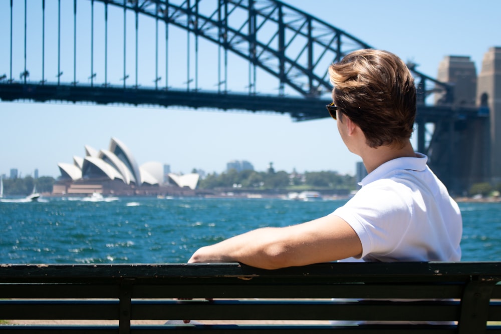 blonde man in white polo shirt sits on bench facing river