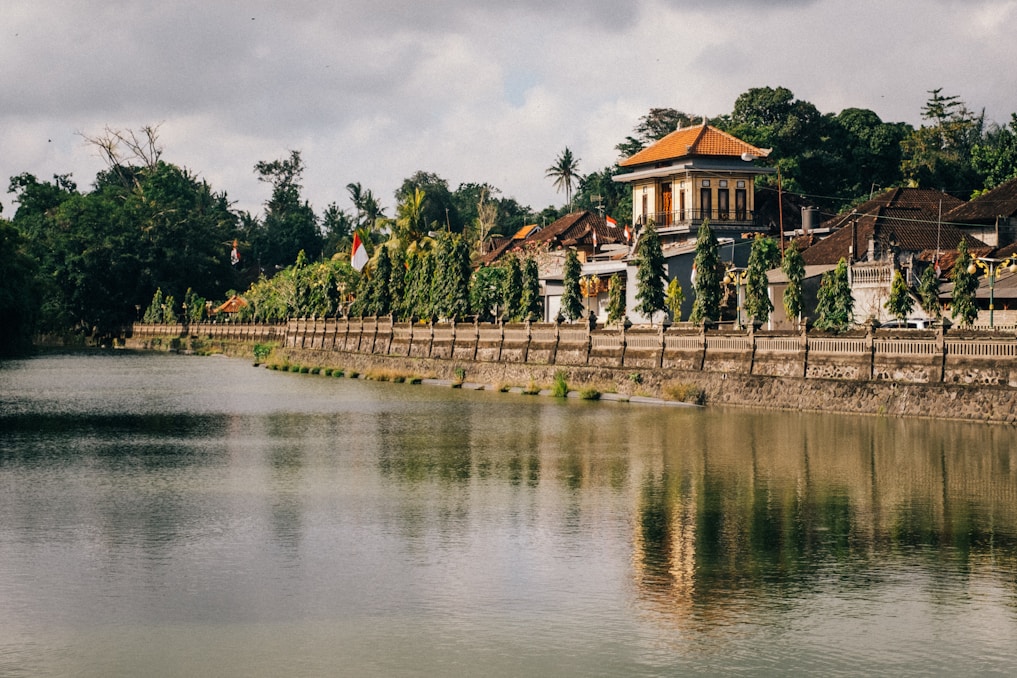 An idyllic riverside scene features a traditional Indonesian building with a red-tiled roof surrounded by lush greenery. The river reflects the structure and surrounding trees, creating a serene, mirrored effect. Flags and decorative elements adorn the area, adding vibrant colors amidst the earthy tones.
