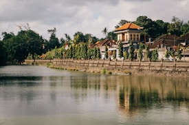 An idyllic riverside scene features a traditional Indonesian building with a red-tiled roof surrounded by lush greenery. The river reflects the structure and surrounding trees, creating a serene, mirrored effect. Flags and decorative elements adorn the area, adding vibrant colors amidst the earthy tones.