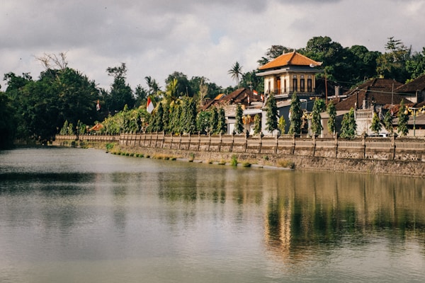 An idyllic riverside scene features a traditional Indonesian building with a red-tiled roof surrounded by lush greenery. The river reflects the structure and surrounding trees, creating a serene, mirrored effect. Flags and decorative elements adorn the area, adding vibrant colors amidst the earthy tones.