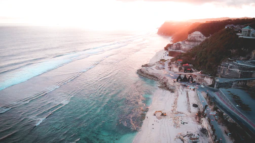 A sweeping aerial view of a coastal village nestled between rugged cliffs and turquoise waters at sunset.