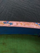 A view of the edge of a boat, featuring a worn surface with faded and peeling paint in shades of blue and yellow. Below the edge, a green surface with visible texture and scratches can be seen. The background is the blurred greyish-green water, indicating the boat is surrounded by water.