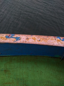 A view of the edge of a boat, featuring a worn surface with faded and peeling paint in shades of blue and yellow. Below the edge, a green surface with visible texture and scratches can be seen. The background is the blurred greyish-green water, indicating the boat is surrounded by water.