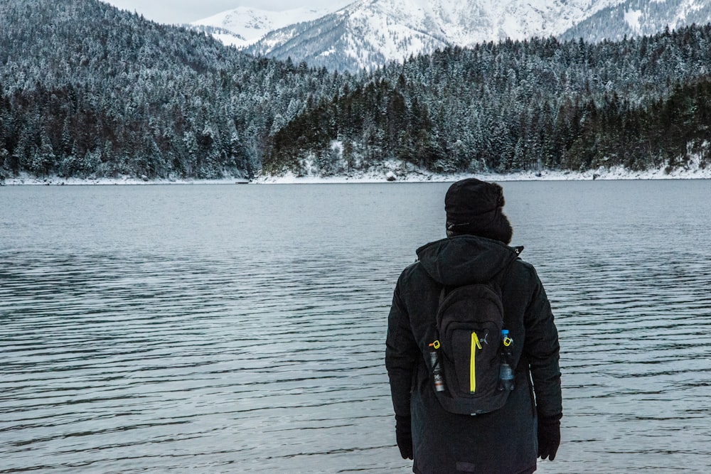 man wearing jacket and backpack facing body of water and mountain