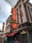 A neon sign reading 'Safari Outfitters LTD' is prominently displayed on the exterior of a building with classic architectural elements, like brickwork and tall windows. Below the sign, the facade includes red awnings and the address '208 Fifth Ave.' Other signage includes 'Brews and Eats' and 'Grill Steakhouse'. People are sitting at outdoor tables, suggesting a lively street scene.