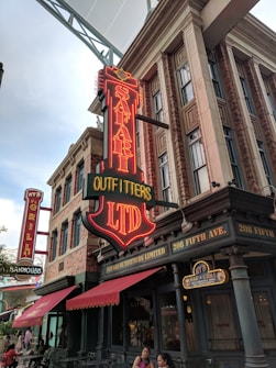 A neon sign reading 'Safari Outfitters LTD' is prominently displayed on the exterior of a building with classic architectural elements, like brickwork and tall windows. Below the sign, the facade includes red awnings and the address '208 Fifth Ave.' Other signage includes 'Brews and Eats' and 'Grill Steakhouse'. People are sitting at outdoor tables, suggesting a lively street scene.