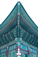 Intricate traditional Asian architectural design featuring vibrant turquoise and teal colors with detailed patterns. The structure exhibits ornate roof eaves, painted beams, and decorative carvings. Symbols and characters are visible along the wooden surfaces, emphasizing cultural artistry.