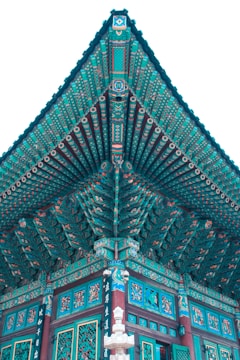 Intricate traditional Asian architectural design featuring vibrant turquoise and teal colors with detailed patterns. The structure exhibits ornate roof eaves, painted beams, and decorative carvings. Symbols and characters are visible along the wooden surfaces, emphasizing cultural artistry.