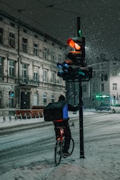A cyclist carrying a delivery box is waiting at a snowy intersection beside a traffic light. The traffic light is illuminated with a red signal, and snow is falling heavily, covering the street and surrounding buildings. The scene takes place at night, casting a dim, cold atmosphere.