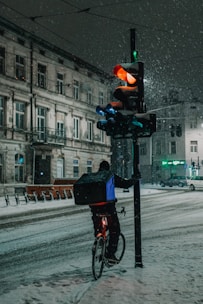 A cyclist carrying a delivery box is waiting at a snowy intersection beside a traffic light. The traffic light is illuminated with a red signal, and snow is falling heavily, covering the street and surrounding buildings. The scene takes place at night, casting a dim, cold atmosphere.