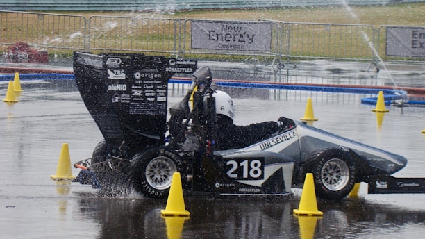 A racing car with the number 218 is navigating a wet track surrounded by yellow traffic cones. The vehicle is sleek with various sponsor logos and has a driver wearing a black suit and white helmet. Water is splashing beneath the tires, indicating recent rain or ongoing drizzle. In the background, banners and metal barriers are visible, suggesting a controlled environment, likely part of a motorsport event.