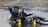 A racing car with the number 218 is navigating a wet track surrounded by yellow traffic cones. The vehicle is sleek with various sponsor logos and has a driver wearing a black suit and white helmet. Water is splashing beneath the tires, indicating recent rain or ongoing drizzle. In the background, banners and metal barriers are visible, suggesting a controlled environment, likely part of a motorsport event.