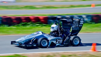 A small, sleek racing car with the number 218 is speeding around a track. The driver is wearing a white helmet, and the car is decorated with various sponsor decals. Orange cones and a barrier made of colorful tires can be seen in the background on the asphalt track.