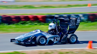 A small, sleek racing car with the number 218 is speeding around a track. The driver is wearing a white helmet, and the car is decorated with various sponsor decals. Orange cones and a barrier made of colorful tires can be seen in the background on the asphalt track.
