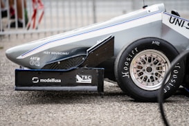 The side view of a race car's front end features sleek aerodynamic lines, a visible tire with 'Hoosier' branding, and multiple sponsor logos on the car body, including 'modelfusa' and 'TRC Aeronautica'. The car is positioned on a coarse, gray textured surface and surrounded by metal fencing in the background.