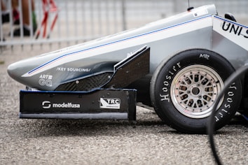 The side view of a race car's front end features sleek aerodynamic lines, a visible tire with 'Hoosier' branding, and multiple sponsor logos on the car body, including 'modelfusa' and 'TRC Aeronautica'. The car is positioned on a coarse, gray textured surface and surrounded by metal fencing in the background.