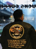 A person is wearing a black jacket with a large emblem on the back featuring an eagle holding a globe and an anchor. The text reads 'United States Marines' with additional text describing the military branches and a quote underneath.