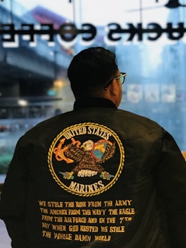A person is wearing a black jacket with a large emblem on the back featuring an eagle holding a globe and an anchor. The text reads 'United States Marines' with additional text describing the military branches and a quote underneath.